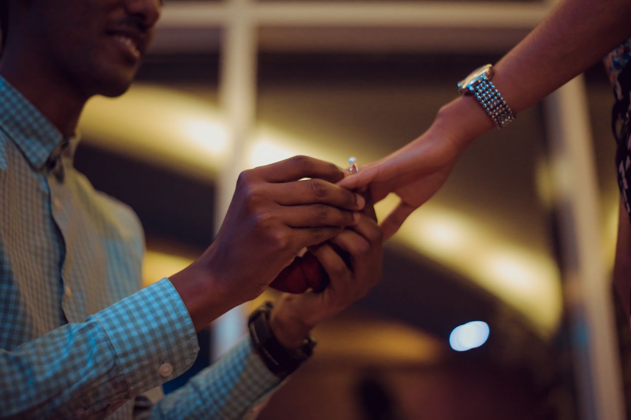 A man gets down on one knee and slips an engagement ring on his partner’s finger in a cityscape