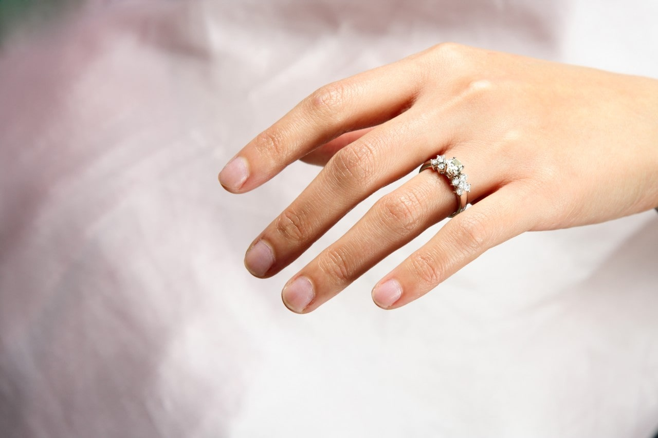 A woman wearing a light pink dress and a side stone engagement ring places her hand in her lap