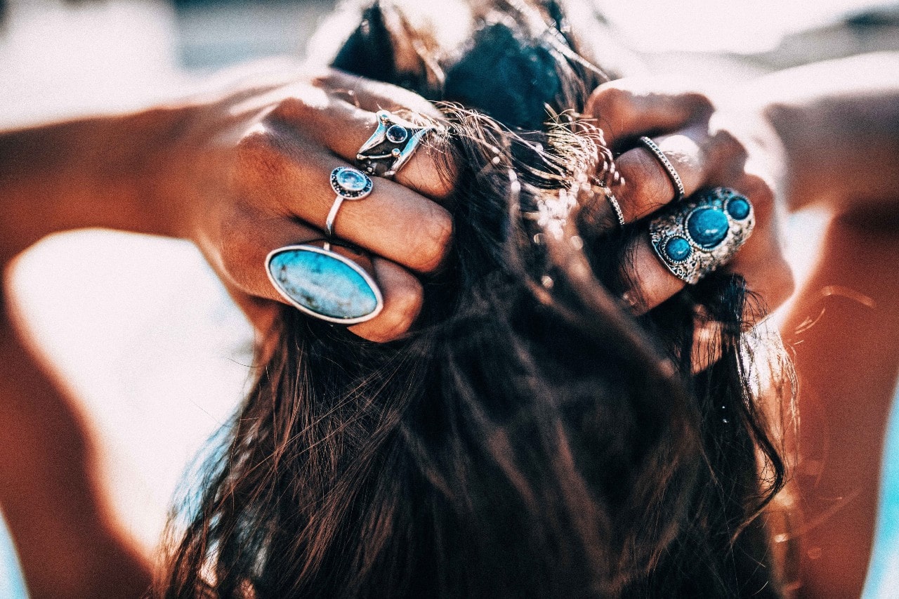 A woman on a beach runs her fingers through her hair while sporting multiple silver and turquoise rings