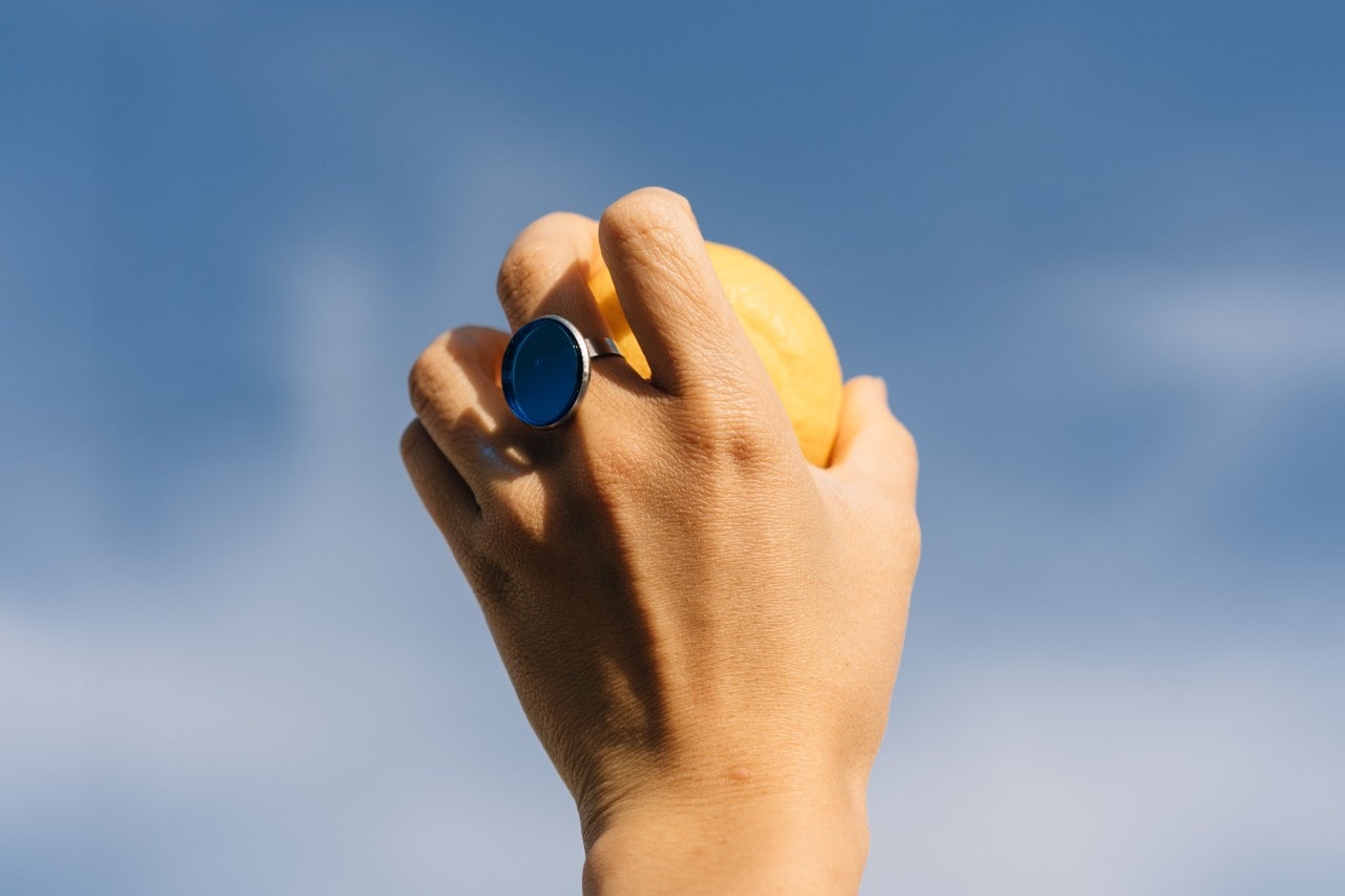 A hand wearing a fashion ring and holding an orange reaches toward the sky