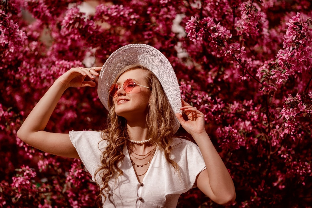 A woman wearing colored sunglasses and a sunhat while standing in front of a flowery bush