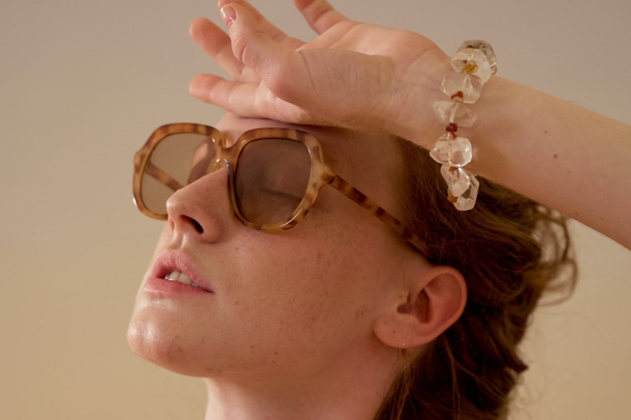 A red-haired woman lounges in front of a beige background, wearing sunglasses, and a chunky bracelet