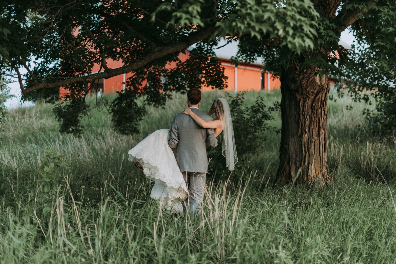 A groom carries his bride through a field and to a barn after their wedding ceremony