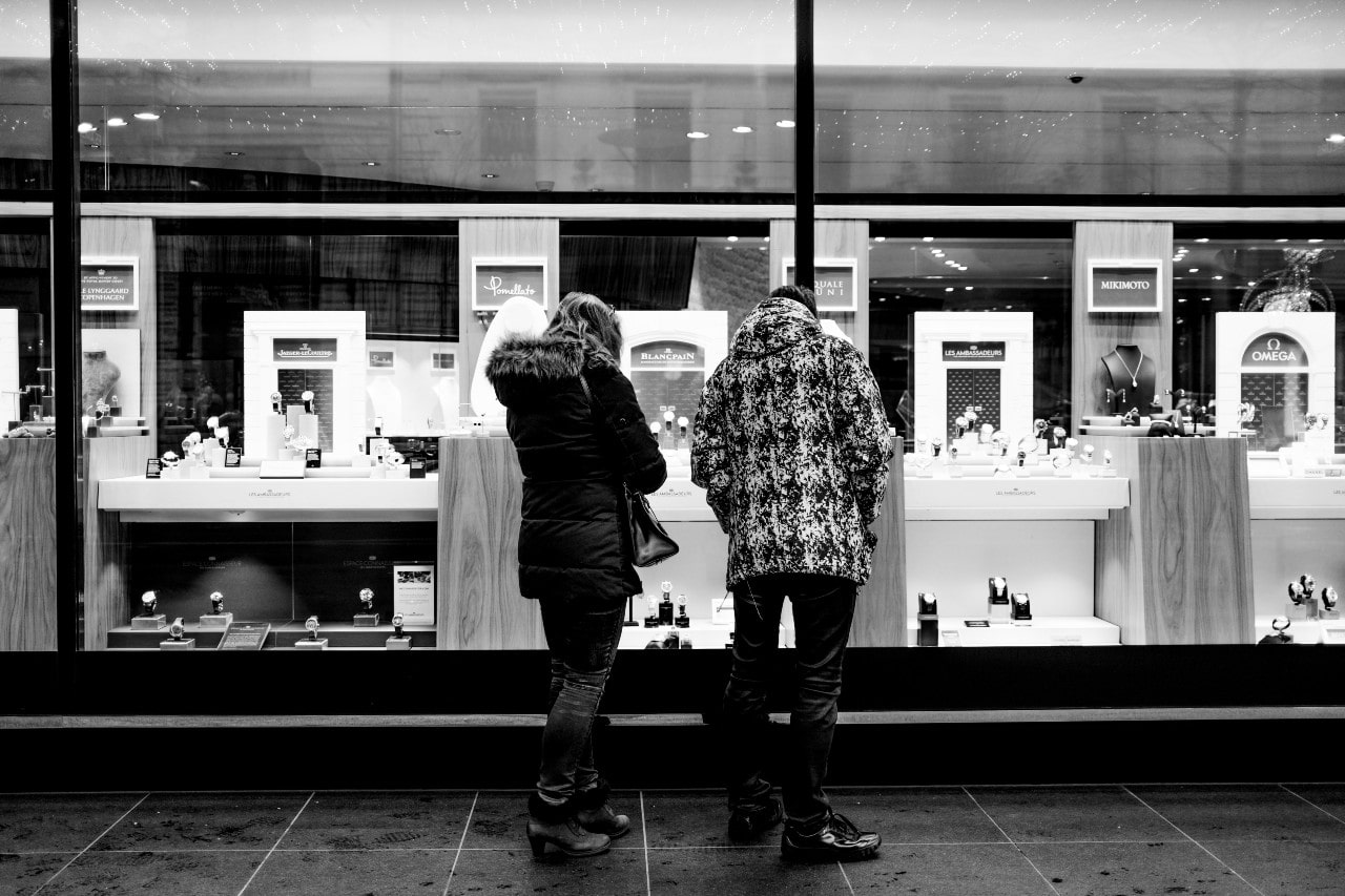 A black-and-white image of a couple standing outside a jewelry store, looking at luxury watches and accessories in the window display.