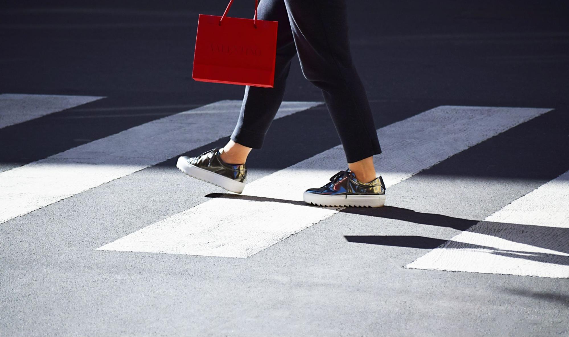A stylish shopper walking across a crosswalk, carrying a red Valentino shopping bag and wearing metallic sneakers.