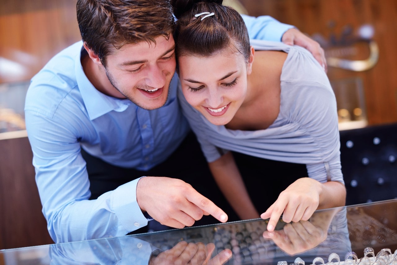 A smiling couple leans over a glass counter in a jewelry store, pointing at sparking engagement rings on display.
