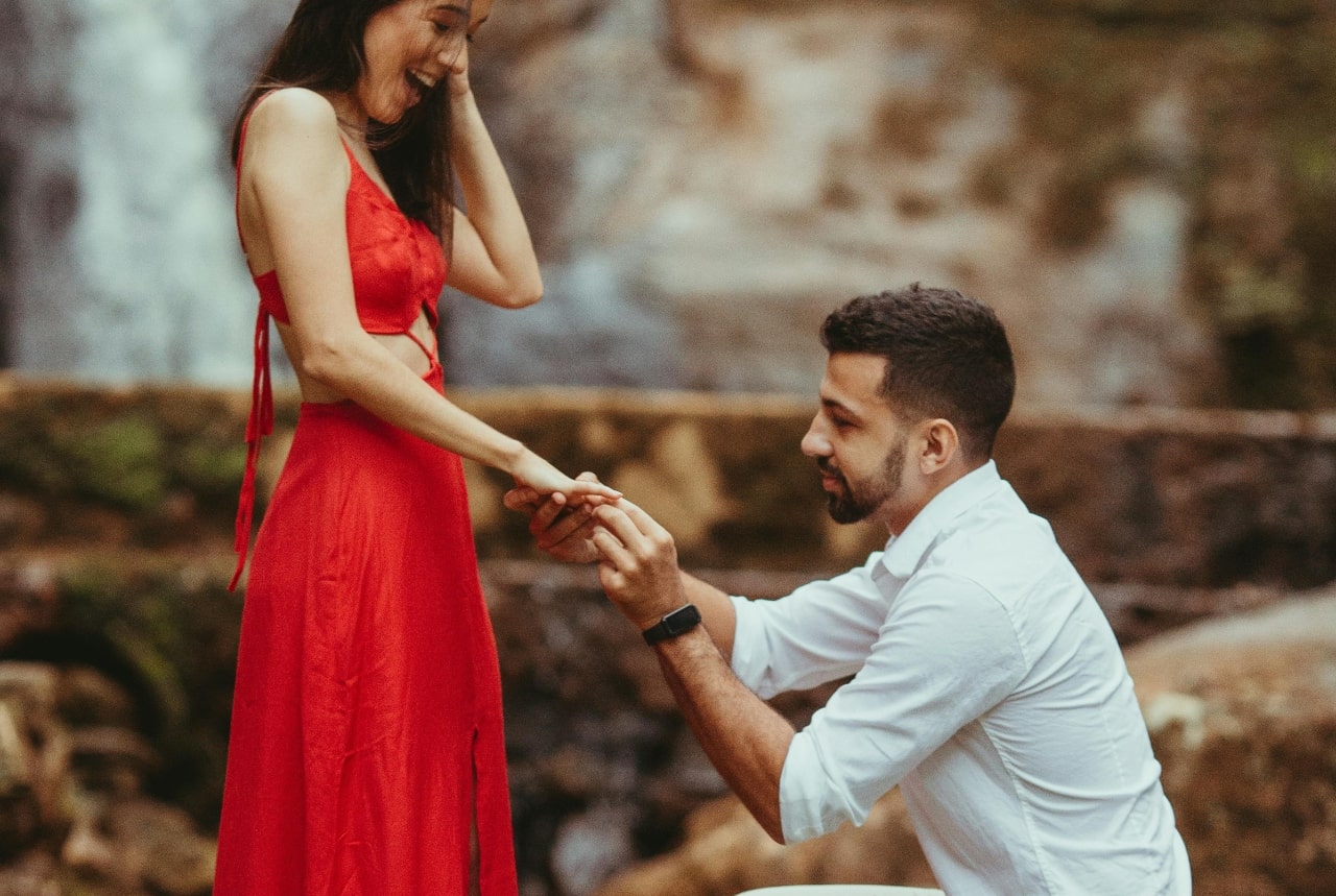 A man kneels, placing an engagement ring on the finger of a surprised woman in a red dress by a waterfall.