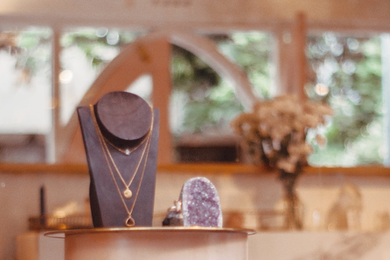A close-up of a jewelry display with layered gold necklaces on a black stand inside a warmly lit boutique.