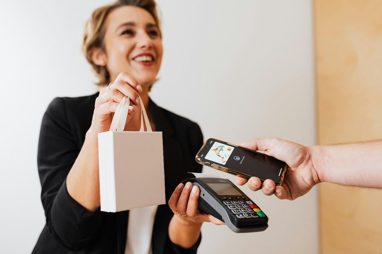A woman in a black blazer is holding a small white shopping bag watches as a person holds a smartphone over a contactless payment terminal