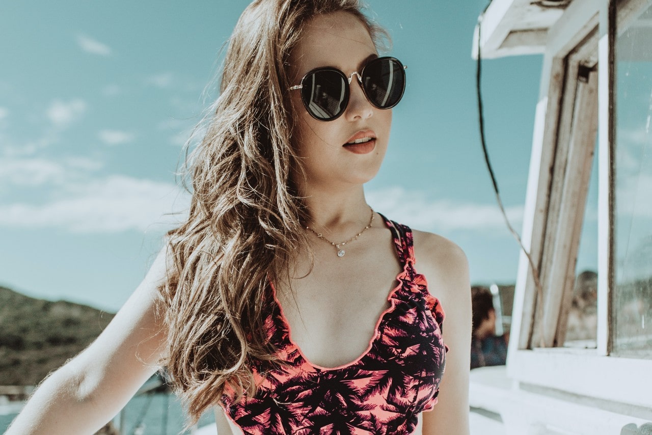 A close up of a woman wearing patterned swimsuits, round sunglasses and a yellow gold necklace, posing on a sunny ship deck