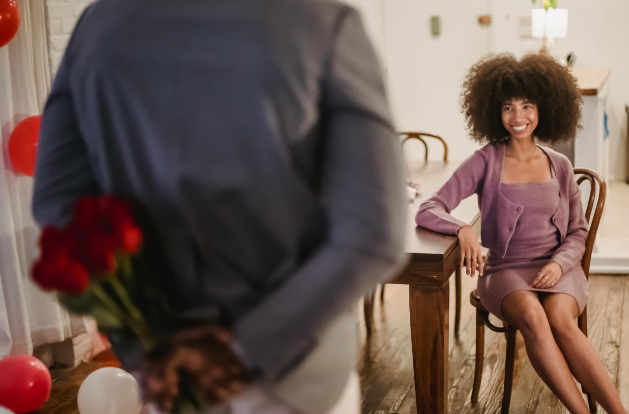 A smiling woman sits at a wooden table while her date hides a bouquet of red roses behind their back.
