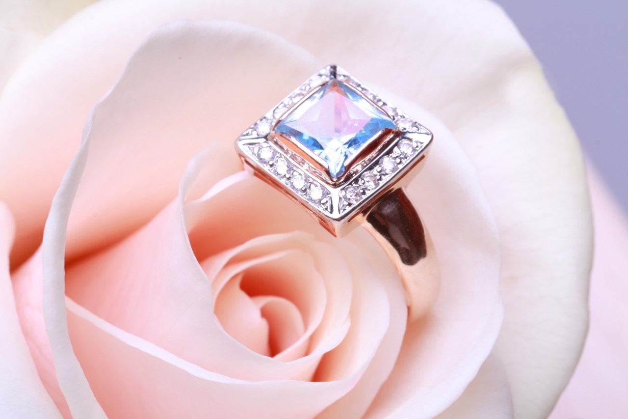 A white gold princess-cut engagement ring atop the soft blush petals of a pale pink rose.