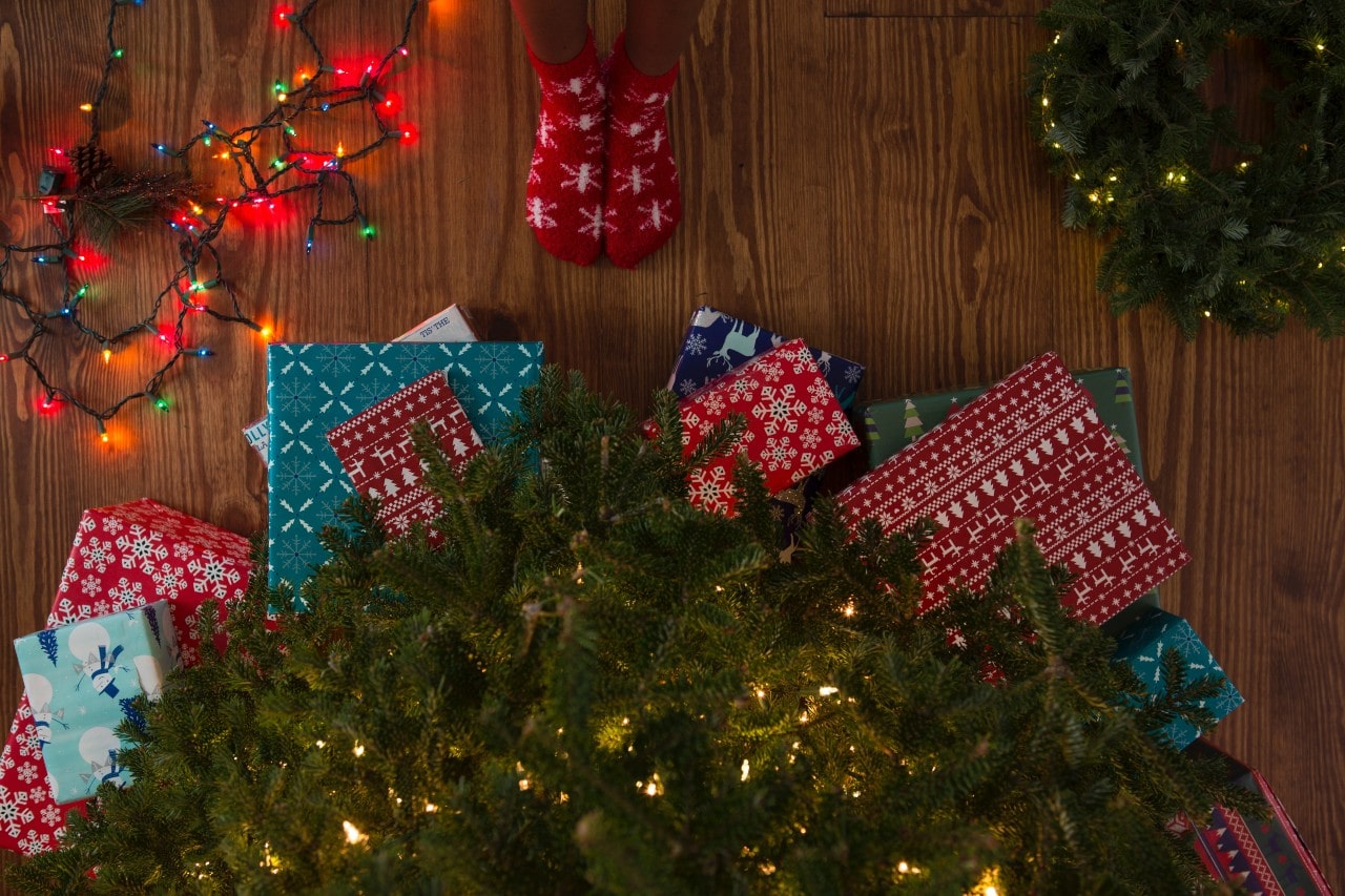 Christmas presents wrapped in festive paper are placed under a decorated tree with colourful string lights and a holiday wreath nearby.