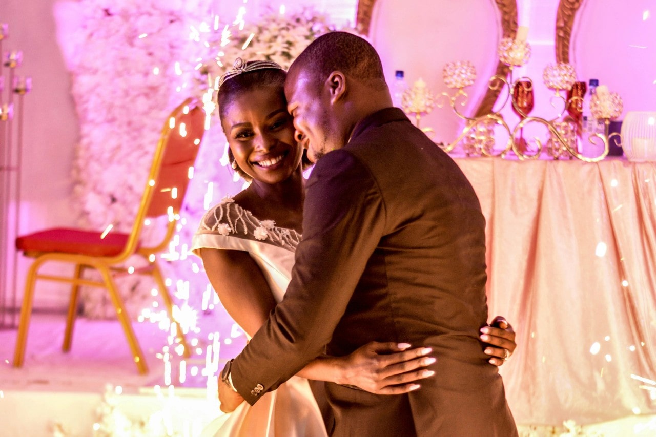 A joyful couple shares an embrace on the dance floor, surrounded by twinkling lights and festive décor, the bride radiant in her white gown and the groom dashing in a brown suit.