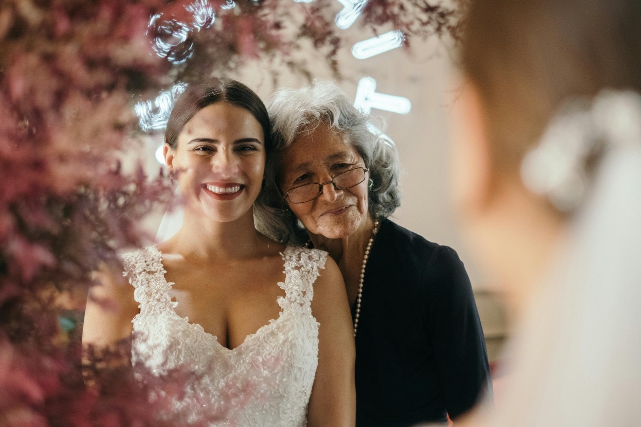 A bride in a white lace gown beams beside an elderly woman wearing glasses, sharing a heartfelt moment amid pink blooms and the soft glow of neon lights.