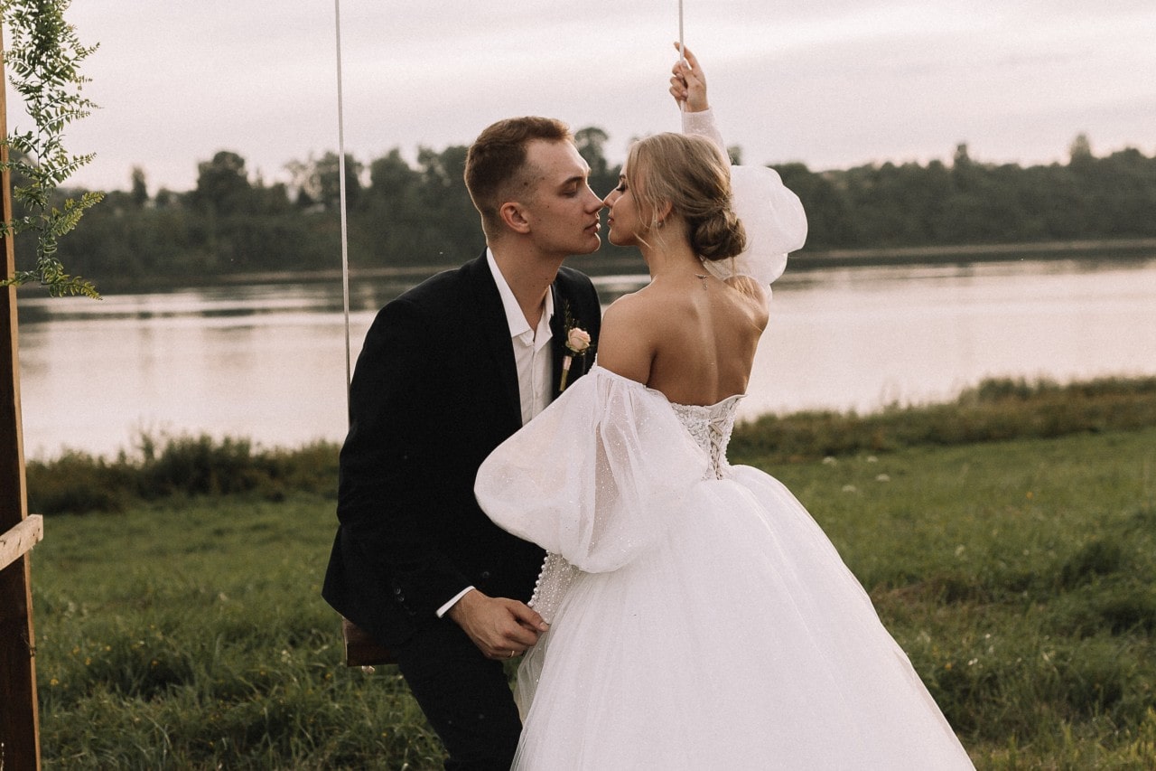 A bride and groom share a tender kiss beside a tranquil lake, the bride’s voluminous white gown flowing gracefully as the groom looks dashing in his black suit.