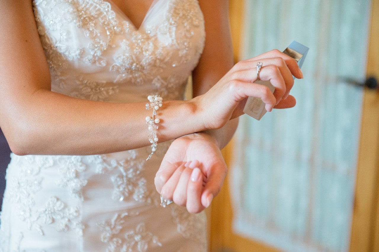 A bride wearing a sparkling bracelet and a ring in an ornate white dress applies perfume on her wrist.