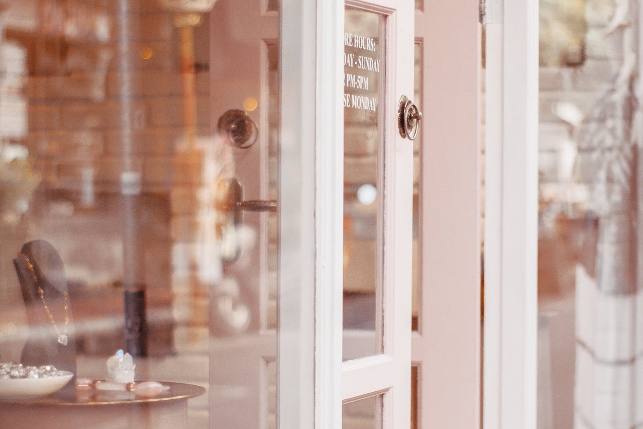 A slightly open pink door with glass panels reveals a jewelry display inside a cozy shop.