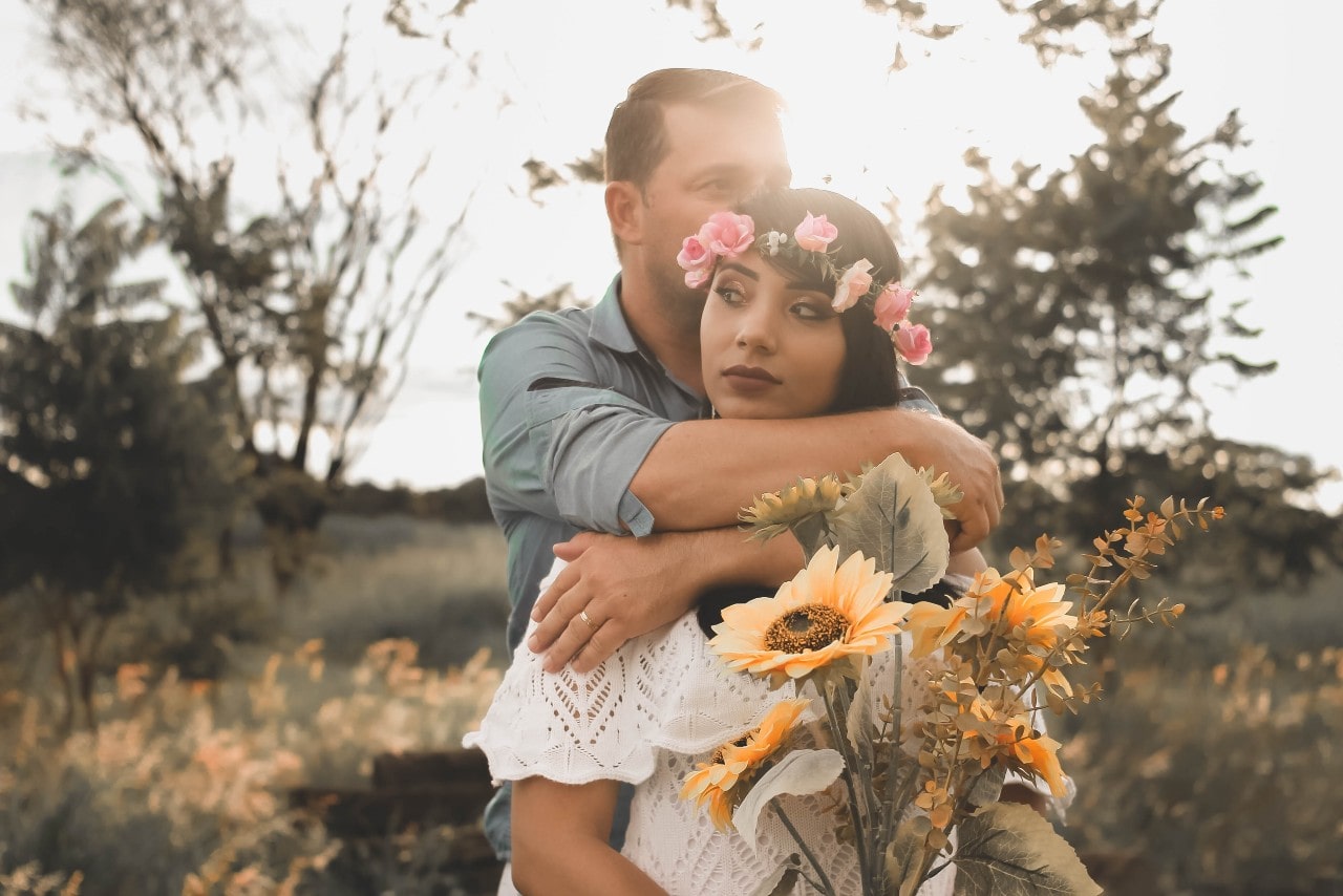 a groom with his arms around his bride, standing outdoors and holding flowers