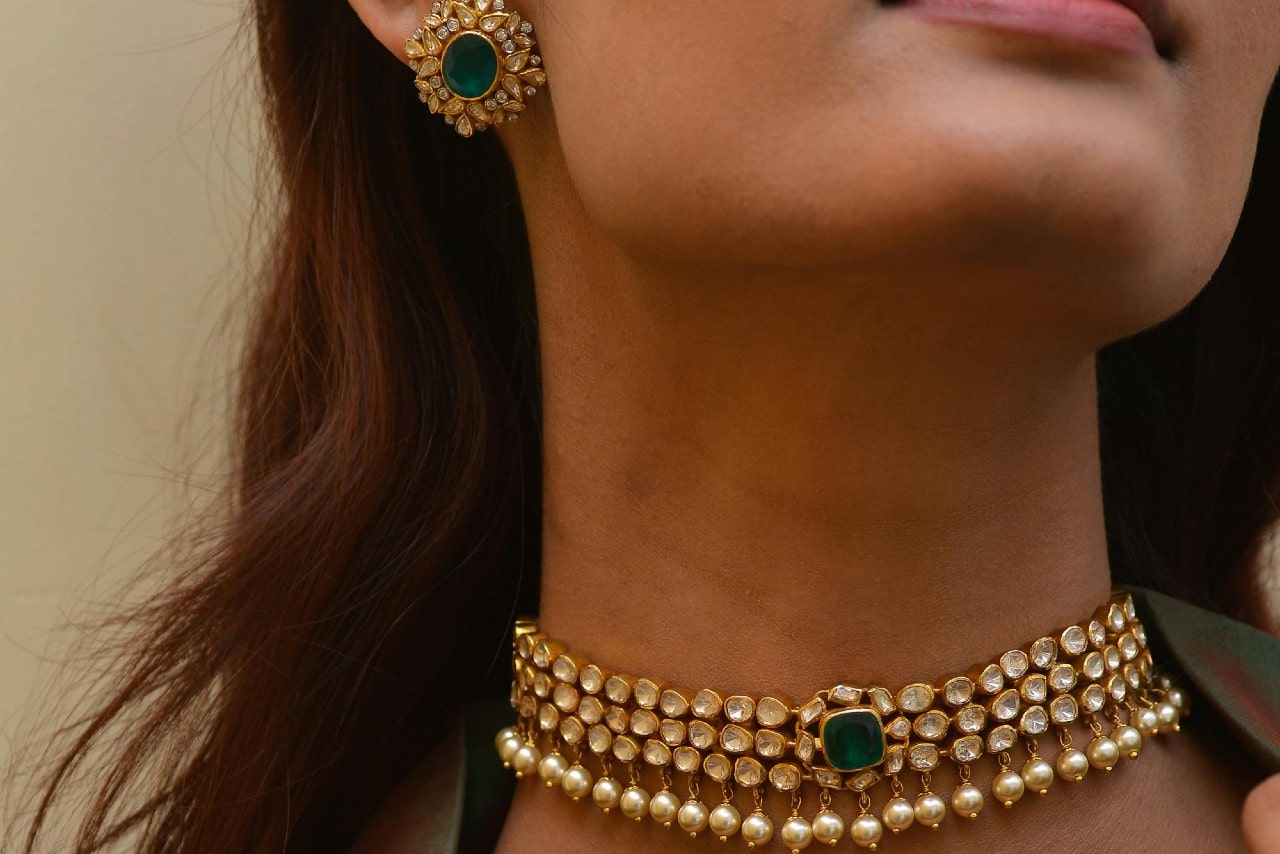 A woman wearing a yellow gold choker adorned with pearls and diamonds, featuring a large green centerpiece, with matching earrings.