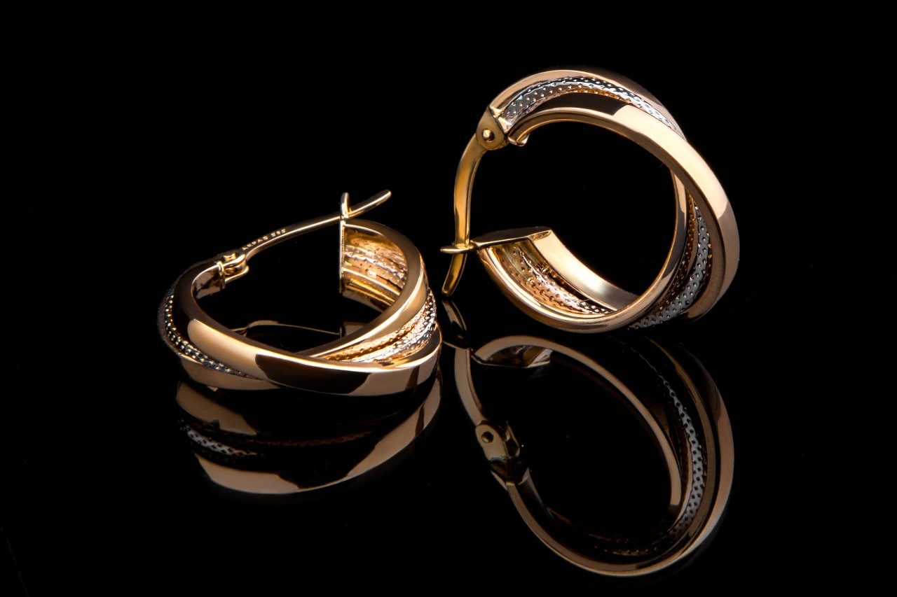 Diamond encrusted gold hoop earrings with intertwined silver bands on a black reflective surface.