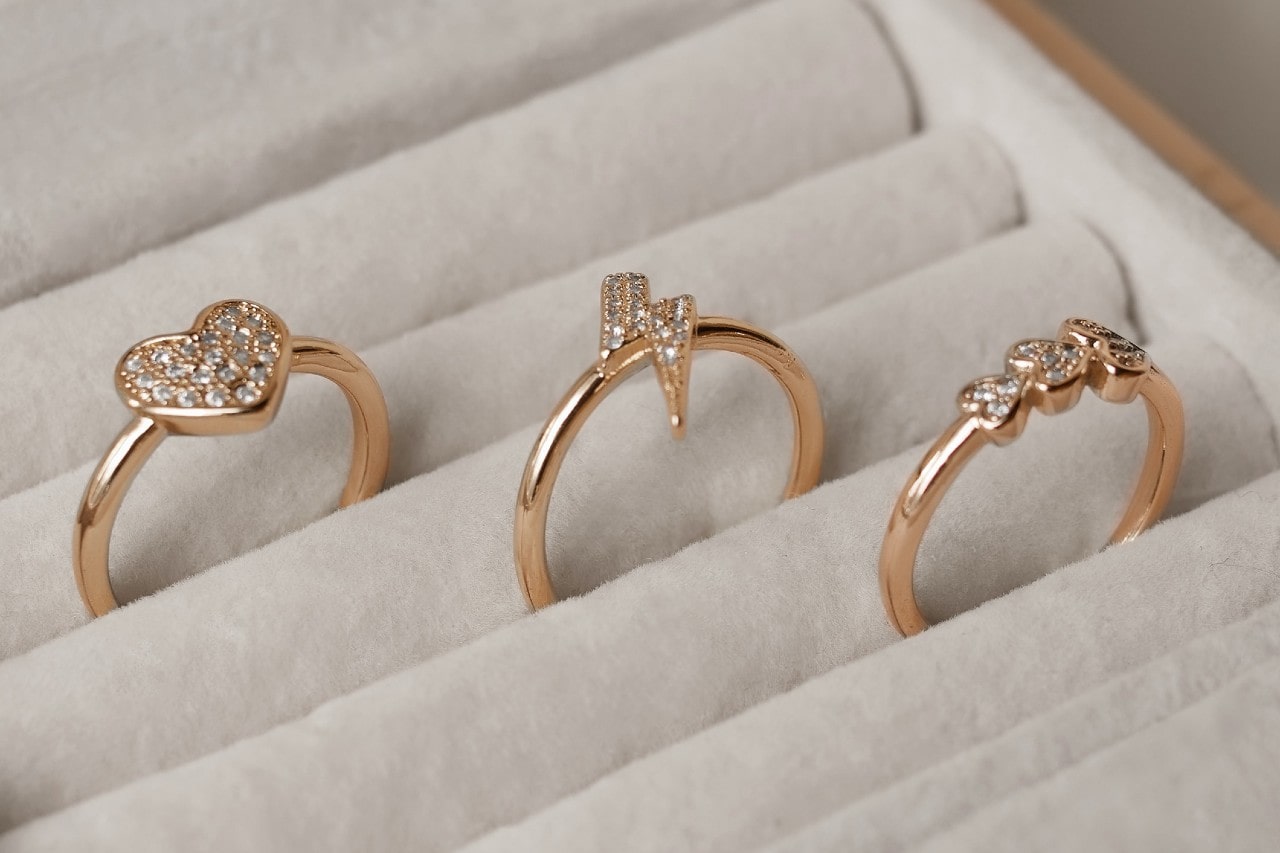 Three gold rings with unique designs rest on a beige ring tray, featuring a heart, lightning bolt, and three stones with pavé settings.