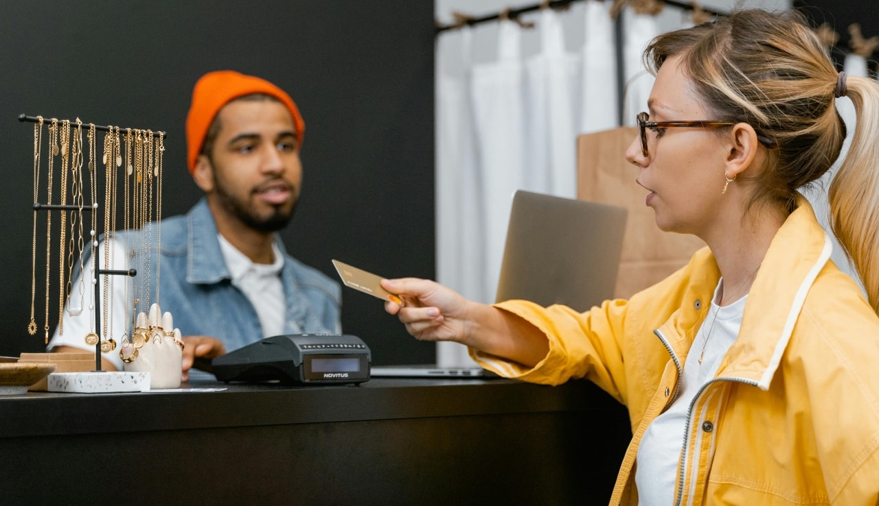 A woman in a yellow jacket hands a credit card to a cashier behind a counter, while he cashier, wearing an orange beanie, stands near a rack of jewelry.