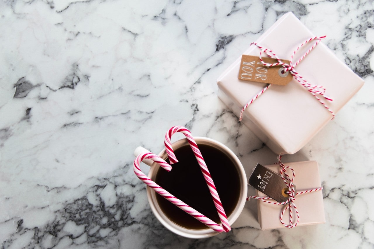 Two candy canes sit in a heart shape on top of a mug beside two wrapped gifts.