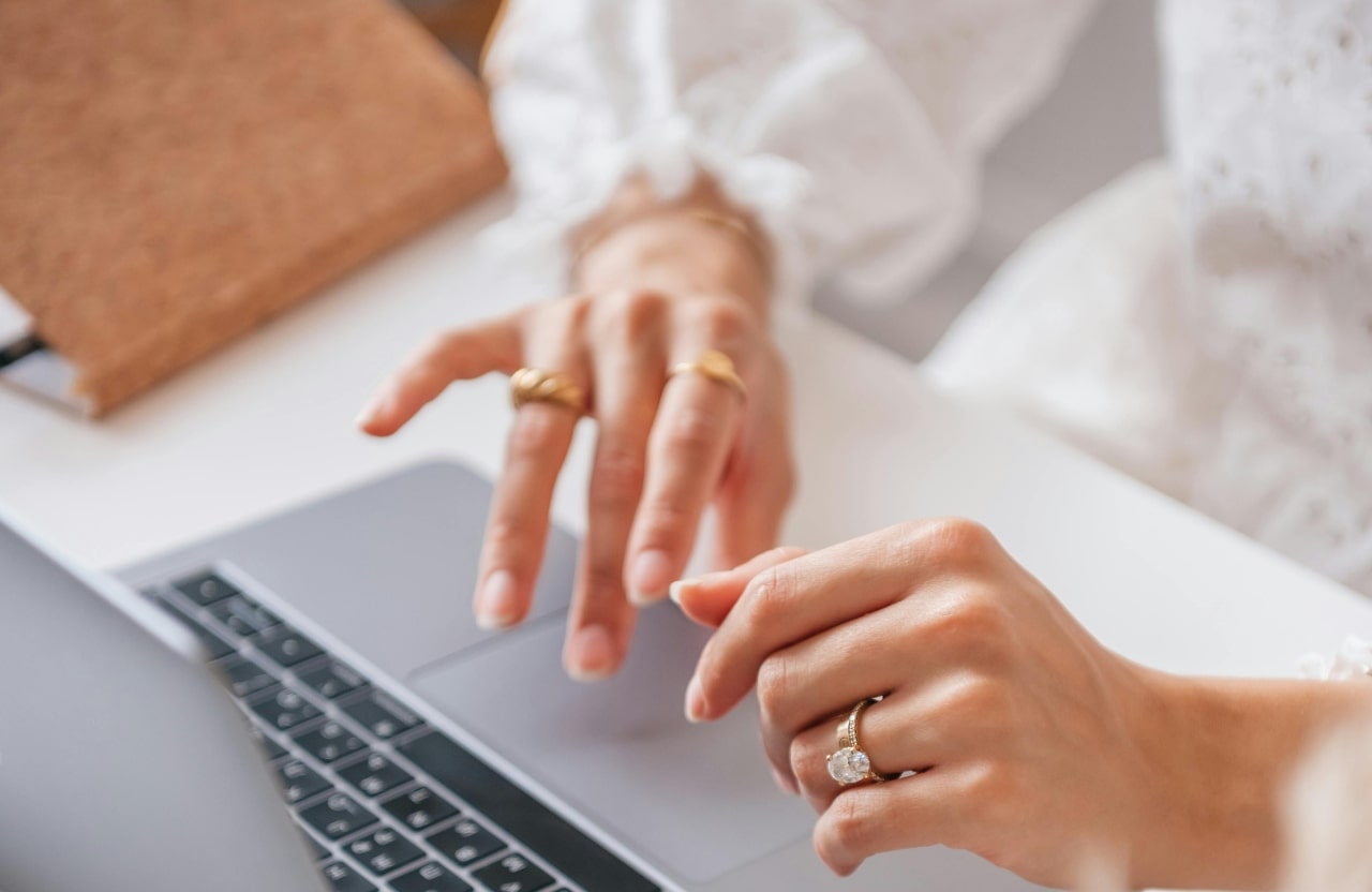A woman working on her laptop with her hands in focus, featuring two yellow gold rings on her right hand, and then a yellow gold oval engagement ring with matching yellow gold thicker wedding band