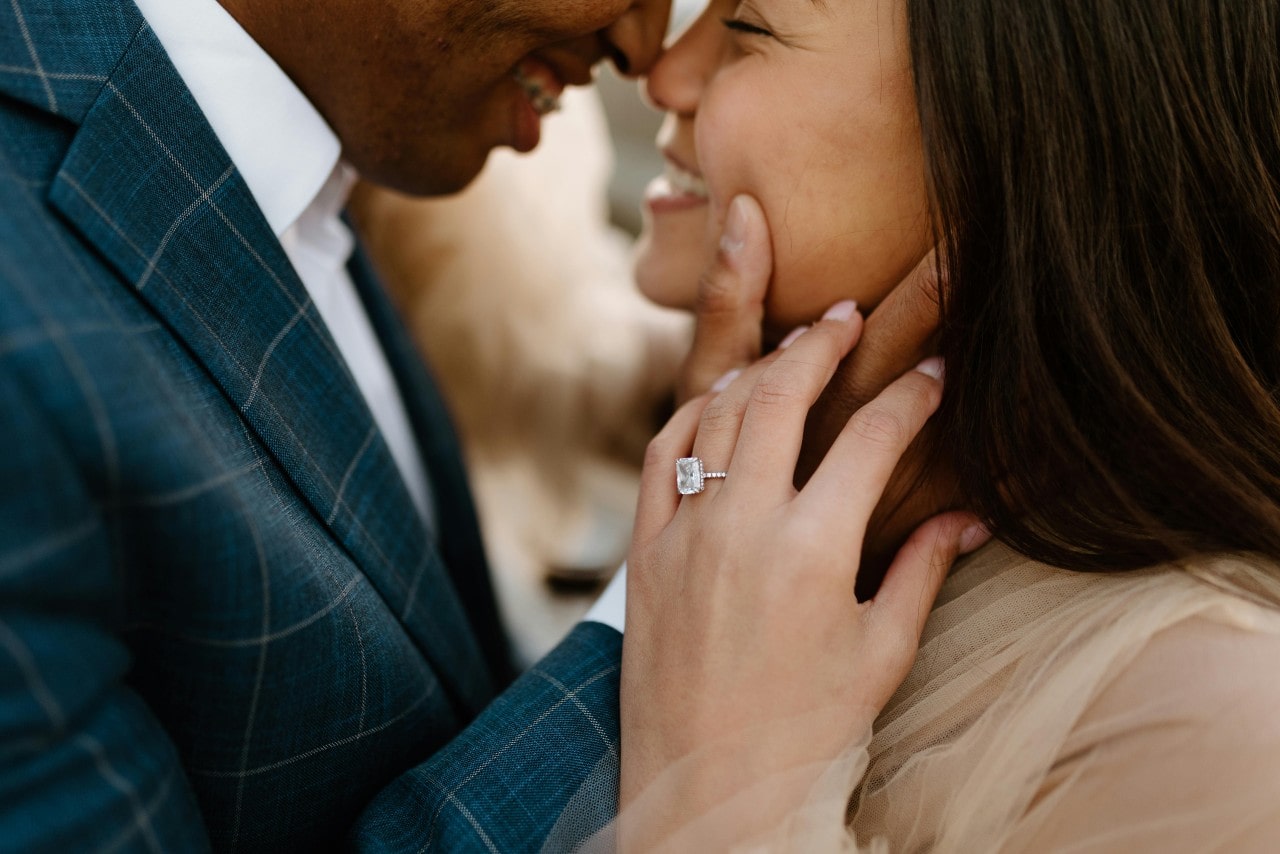A couple about to kiss, and focus is on the radiant cut hidden halo ring with diamonds along the band of the white gold ring as well