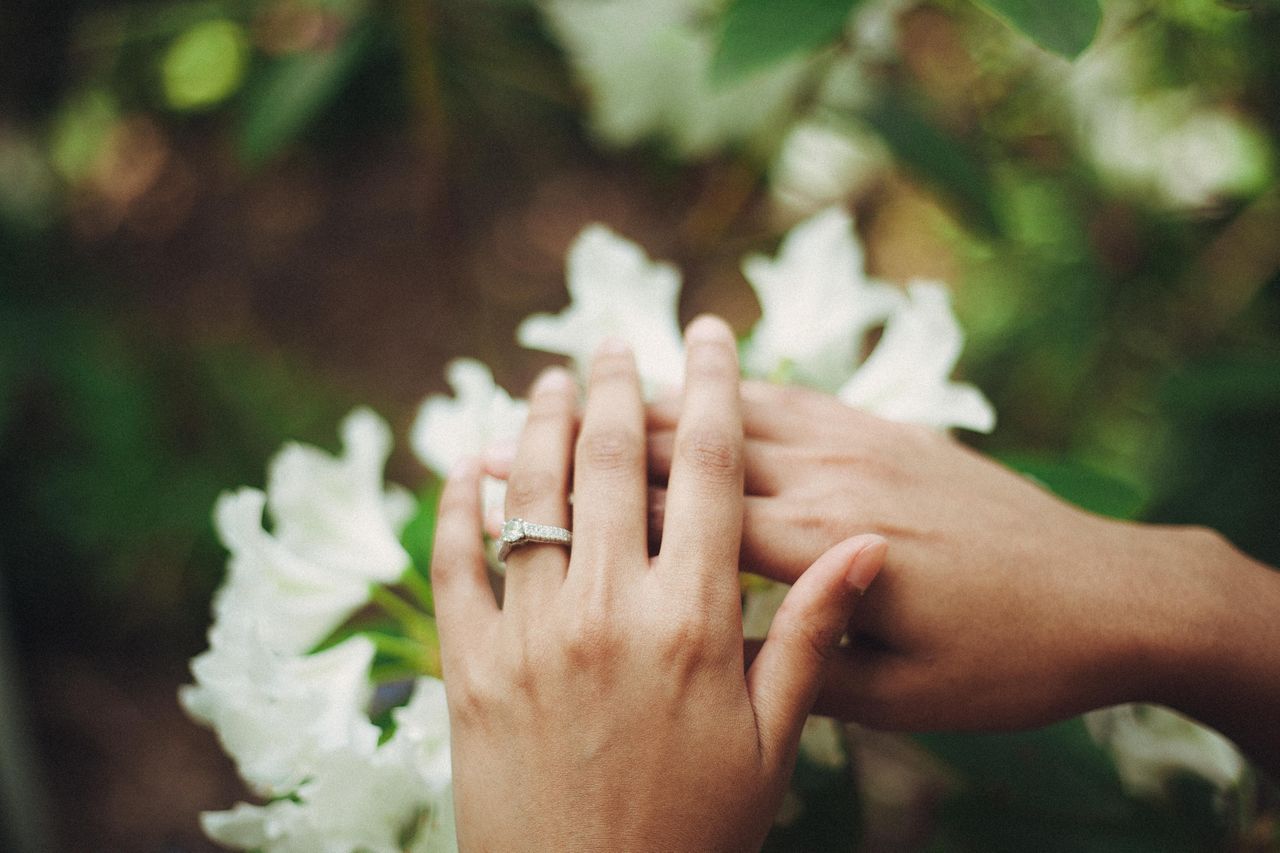 Close up of the couple's hands gently touching white flowers, one adorned with a sparkling ring, surrounded by lush green leaves.