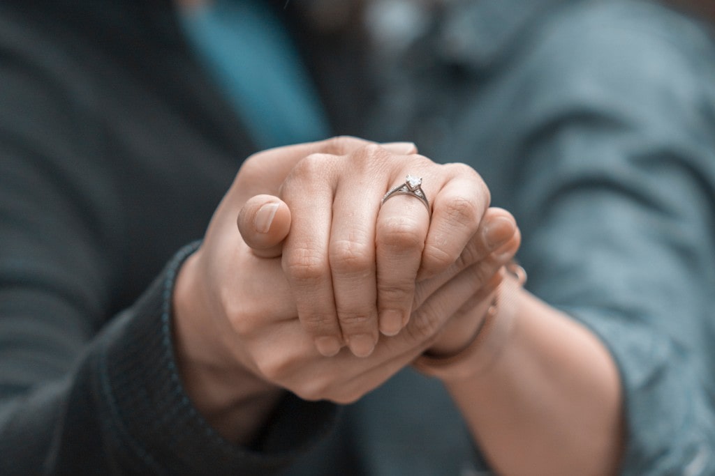 Close up of the couple's hands holding gently, with a strong focus on the beautiful solitaire diamond engagement ring of the woman.