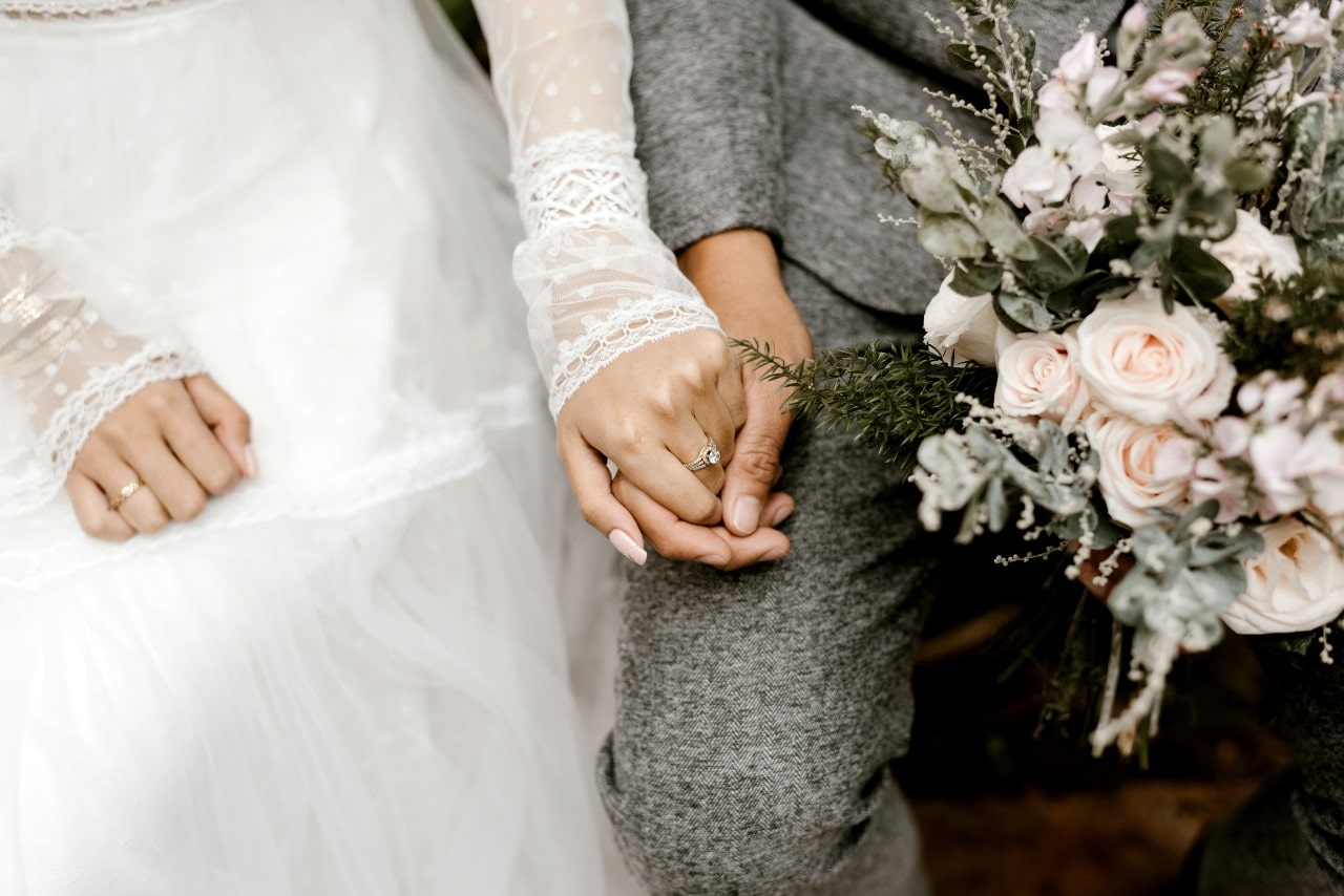 Close-up of a bride and groom's hands intertwined, she in a white lace dress, flaunting her engagement ring and he in a grey suit, holding a bouquet of pastel roses.