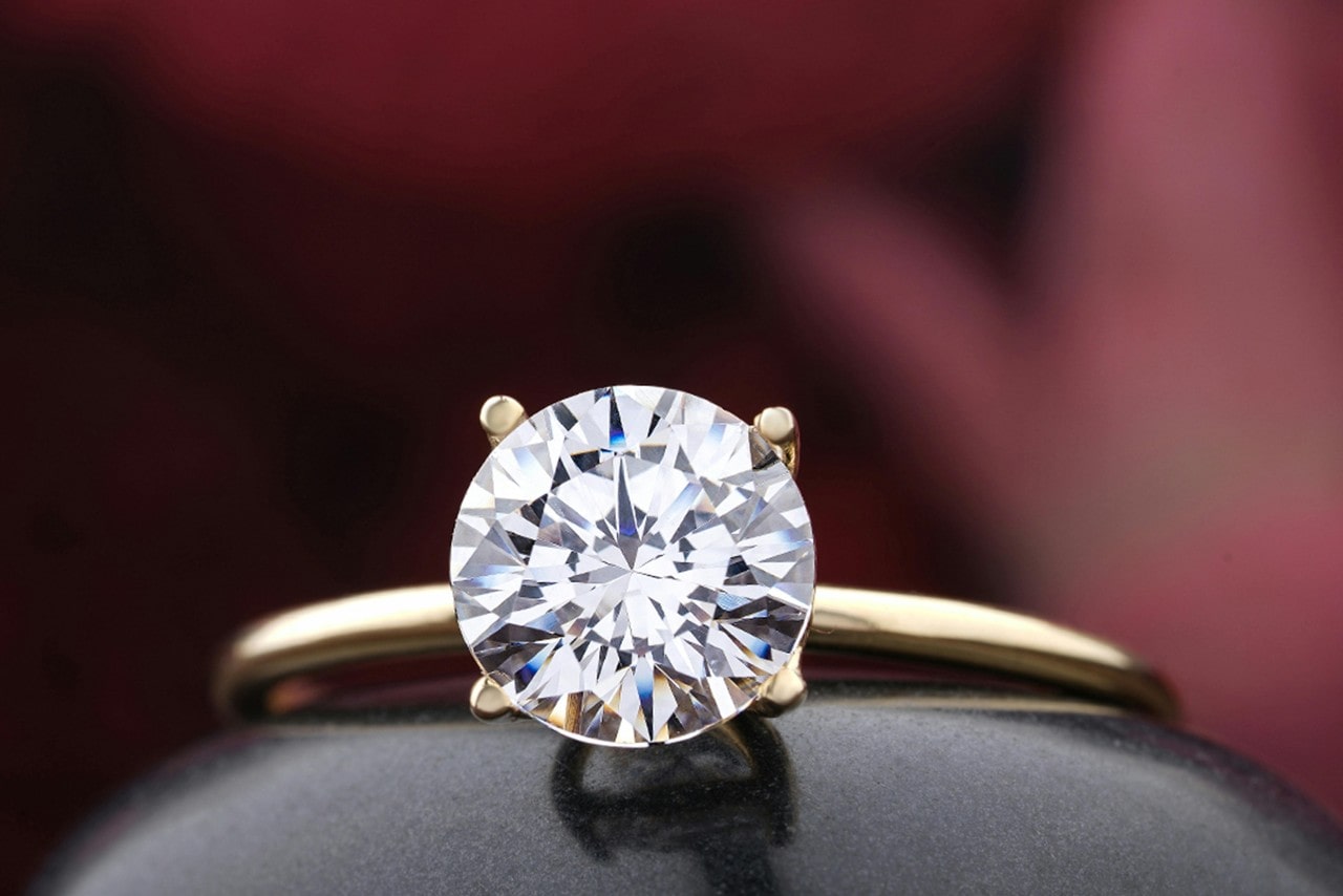 A close up of a round cut yellow gold thin solitaire engagement ring against a blurry red background. A close up of a round cut yellow gold thin solitaire engagement ring against a blurry red background.
