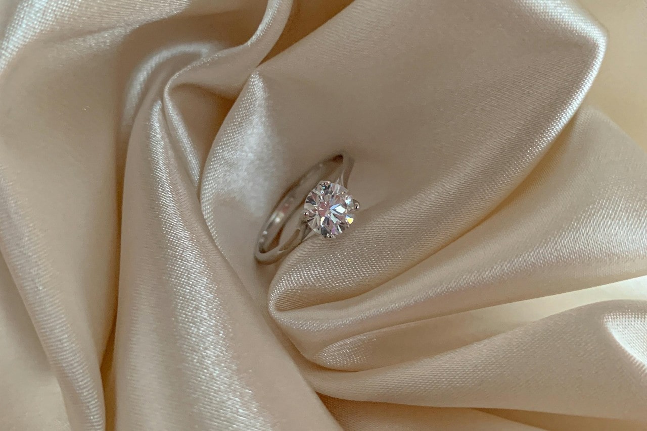 A close up of a white gold round cut diamond engagement ring on a white atlas fabric. A close up of a white gold round cut diamond engagement ring on a white atlas fabric.