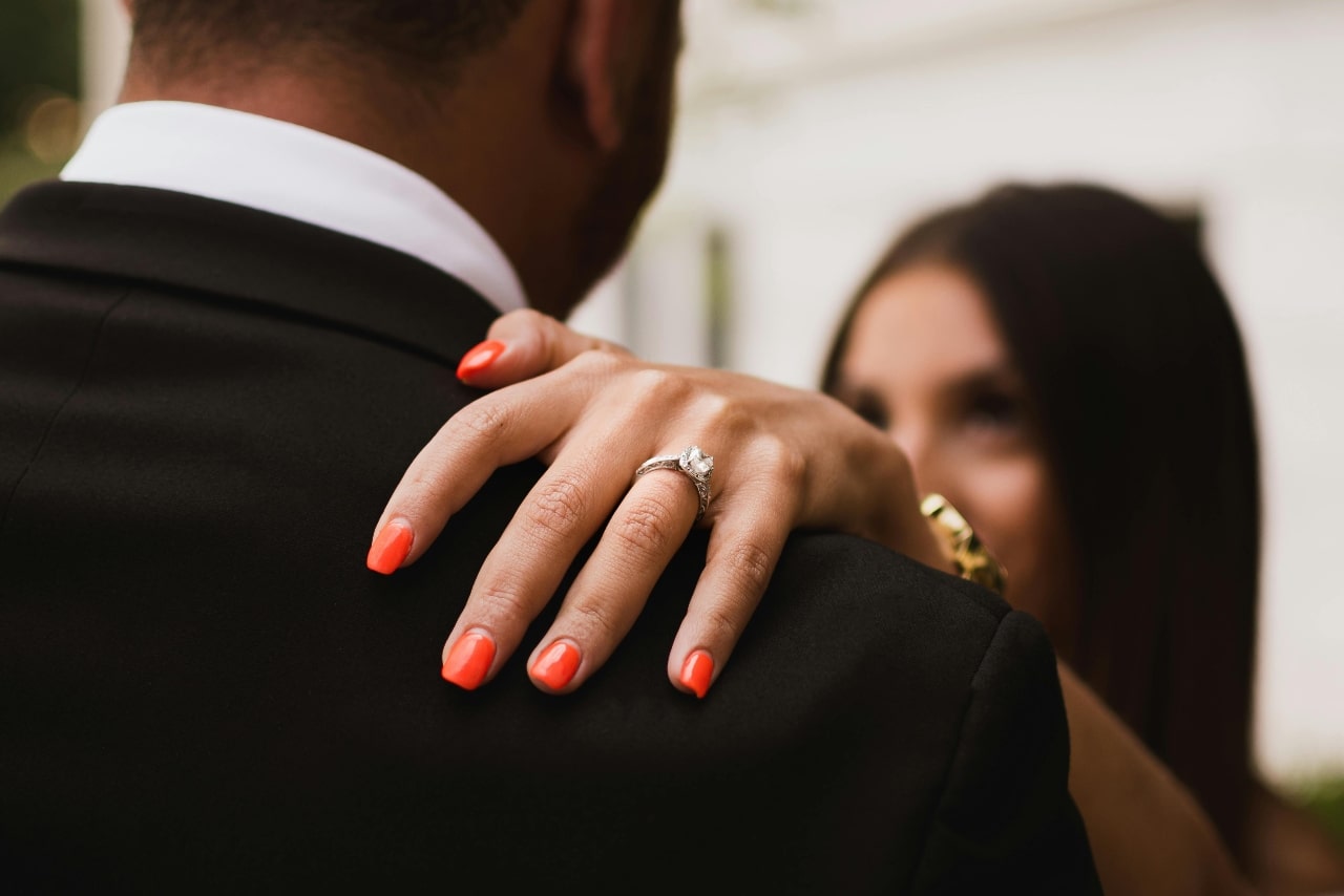 A close-up of a woman’s hand with red manicure and diamond engagement ring, resting on a man's shoulder. A close-up of a woman’s hand with red manicure and diamond engagement ring, resting on a man's shoulder.