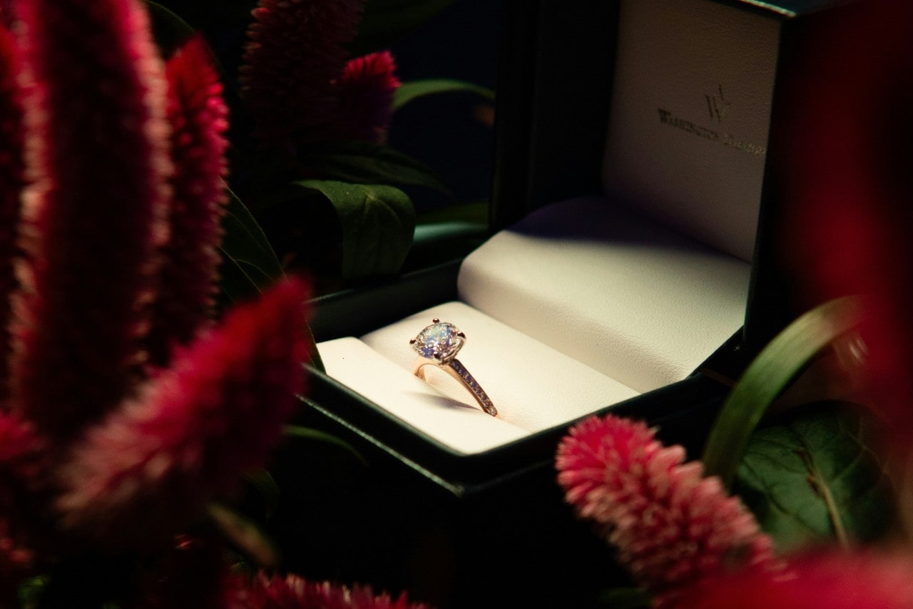 A sidestone yellow gold engagement ring in a box surrounded by pink flowers and leaves. A sidestone yellow gold engagement ring in a box surrounded by pink flowers and leaves.