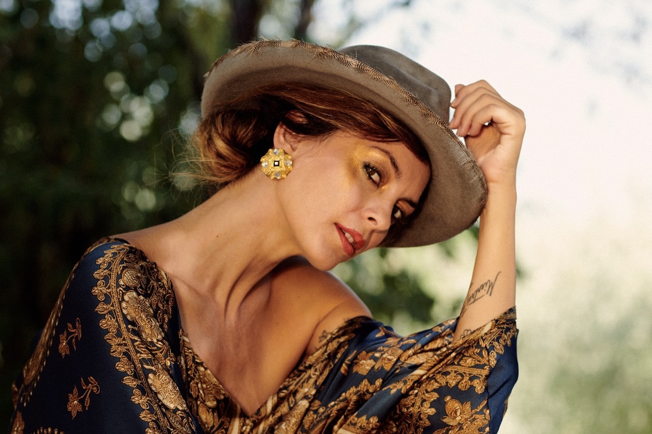 Woman in a wide-brimmed hat, wearing golden earrings and an ornate floral dress, tilts her head.
