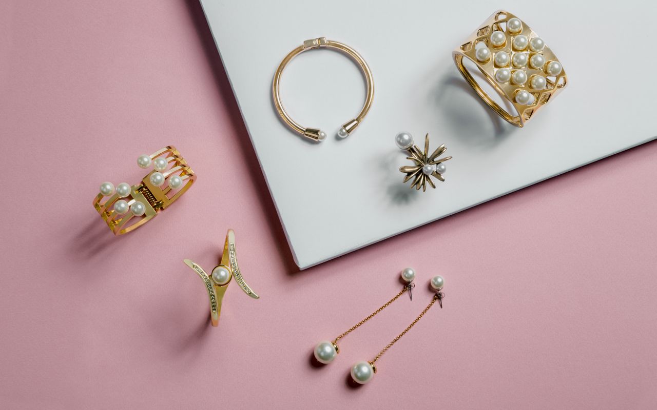 A set of gold and pearl jewelry on a pink surface, including bracelets, earrings, and a ring, laid out on a white square.