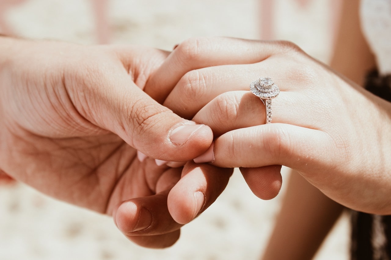 A woman’s hand held by a man’s hand, with emphasis on a stunning diamond halo engagement ring.