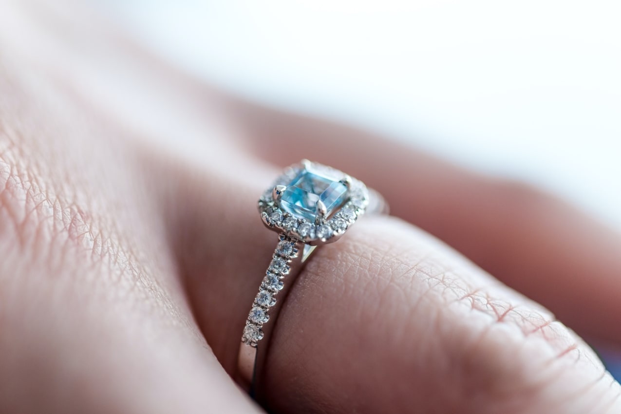 A close up of a gemstone halo engagement ring on a finger.