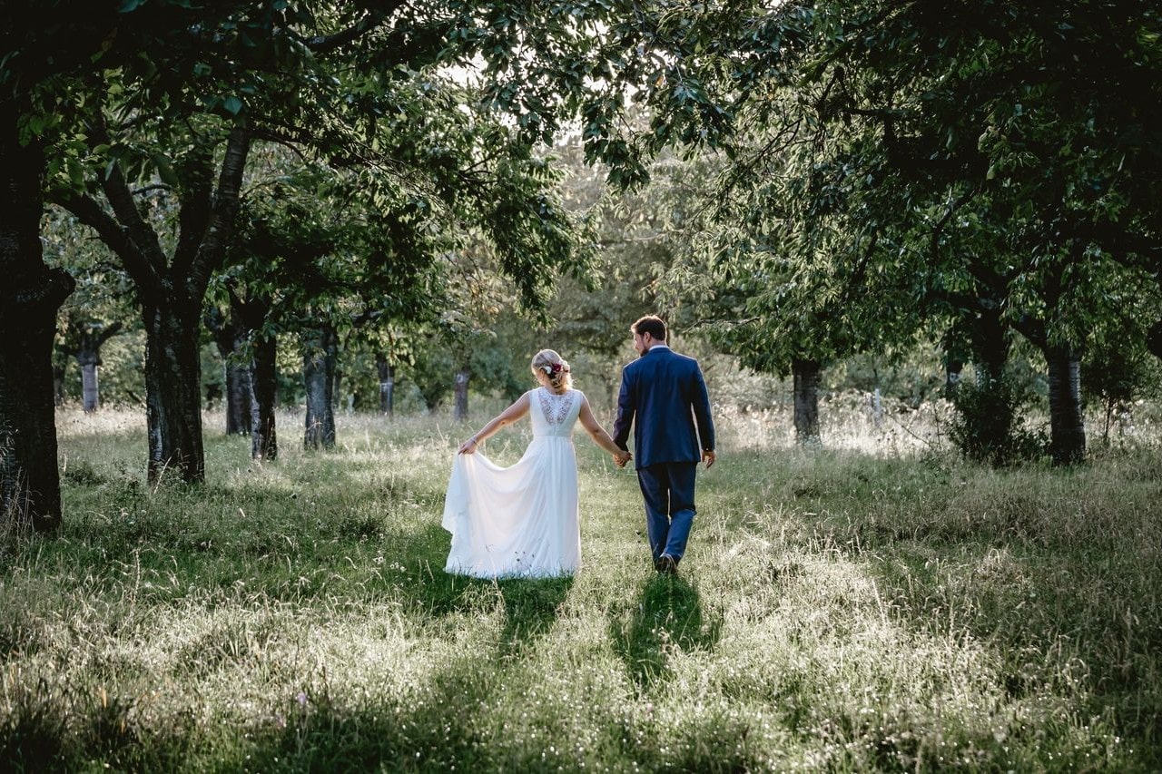 A newly-wed couple walks through a field while holding hands.