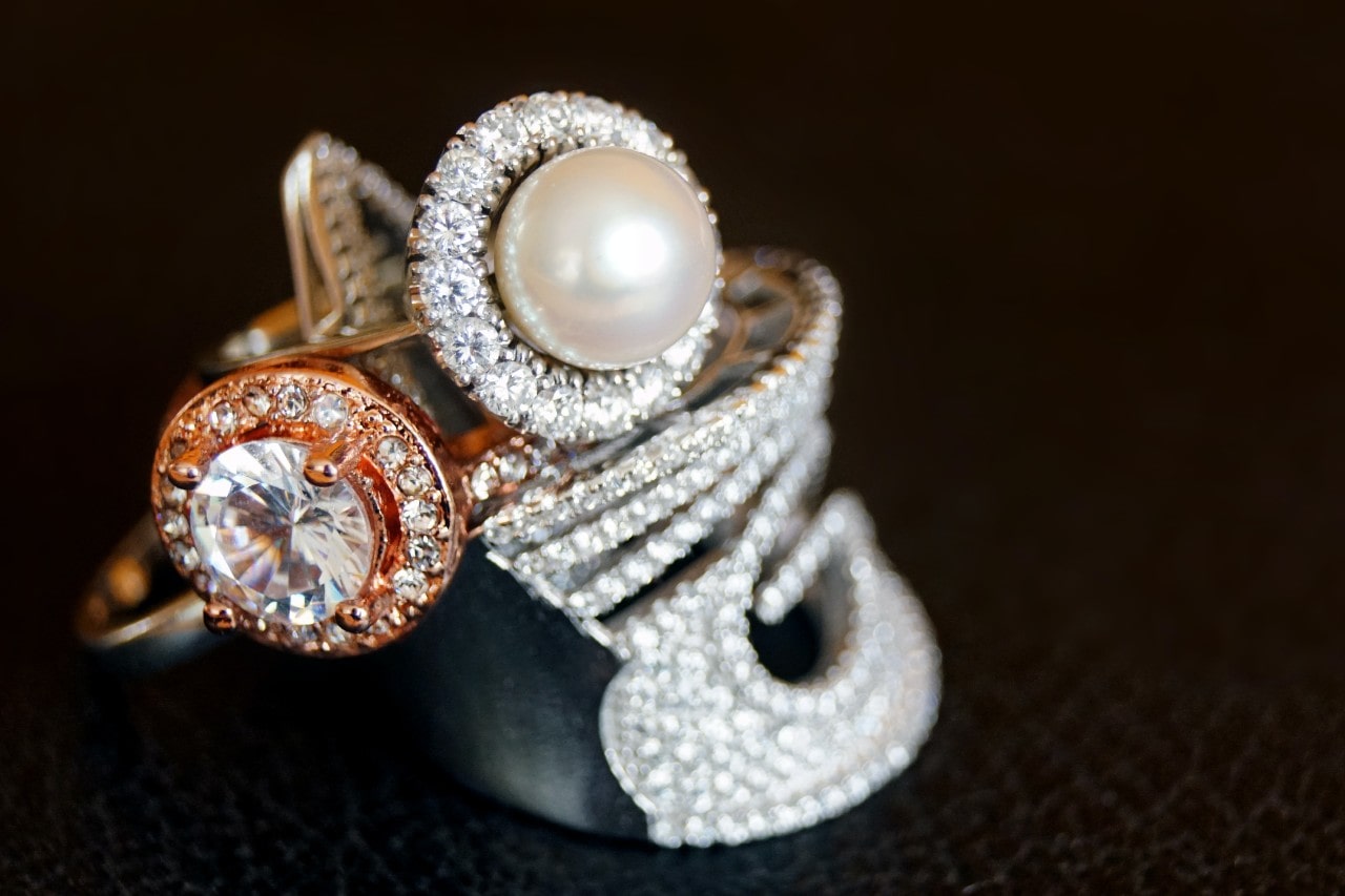 A close-up of two rings stacked together, one with a large white pearl surrounded by small diamonds, and the other with round diamond in a rose gold setting, both resting on a dark, textured surface. A close-up of two rings stacked together, one with a large white pearl surrounded by small diamonds, and the other with round diamond in a rose gold setting, both resting on a dark, textured surface.