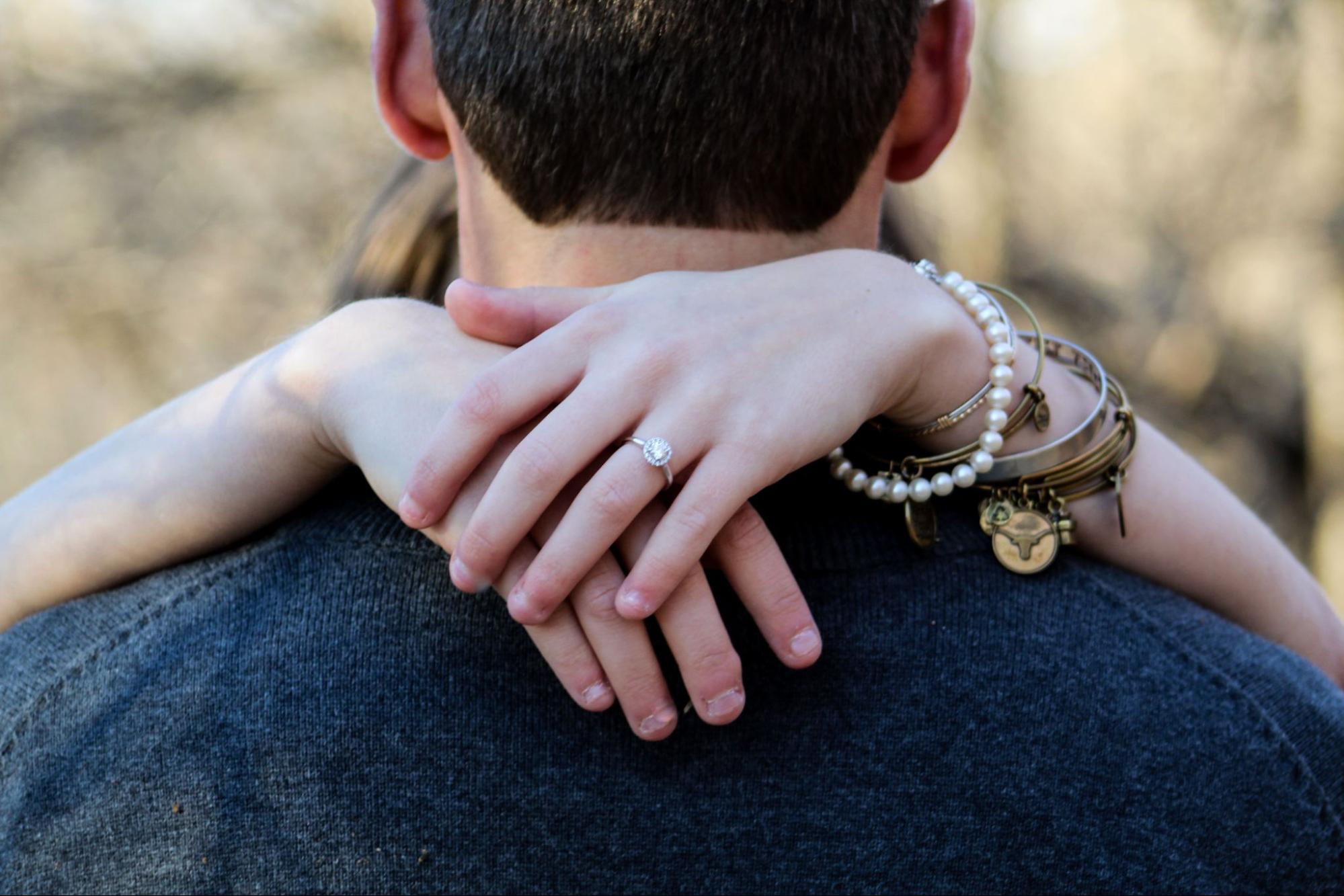A close up of a woman's hands resting on a man’s shoulder, showcasing her pearl bracelet, cushion cut diamond engagement ring and various other bracelets. A close up of a woman's hands resting on a man’s shoulder, showcasing her pearl bracelet, cushion cut diamond engagement ring and various other bracelets.