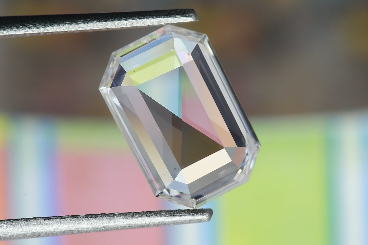 A precision-cut, emerald-shaped gemstone reflects pastel hues while held by tweezers.