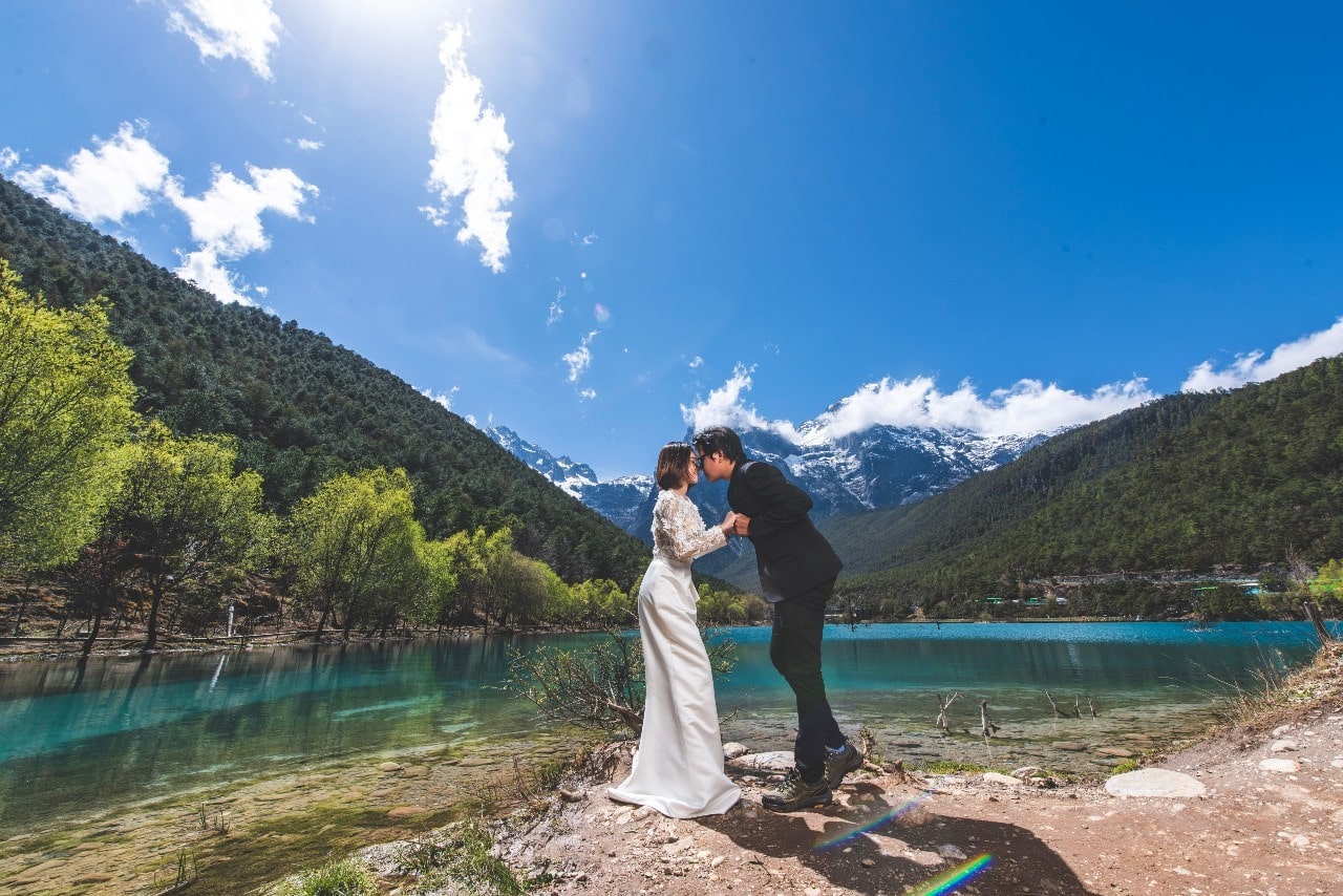 A bride and groom pose to kiss by a pond in the mountains.