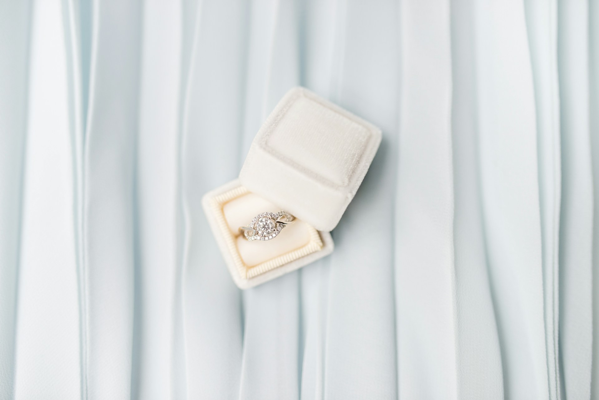 A diamond halo engagement ring in a white box on a white pleated fabric.