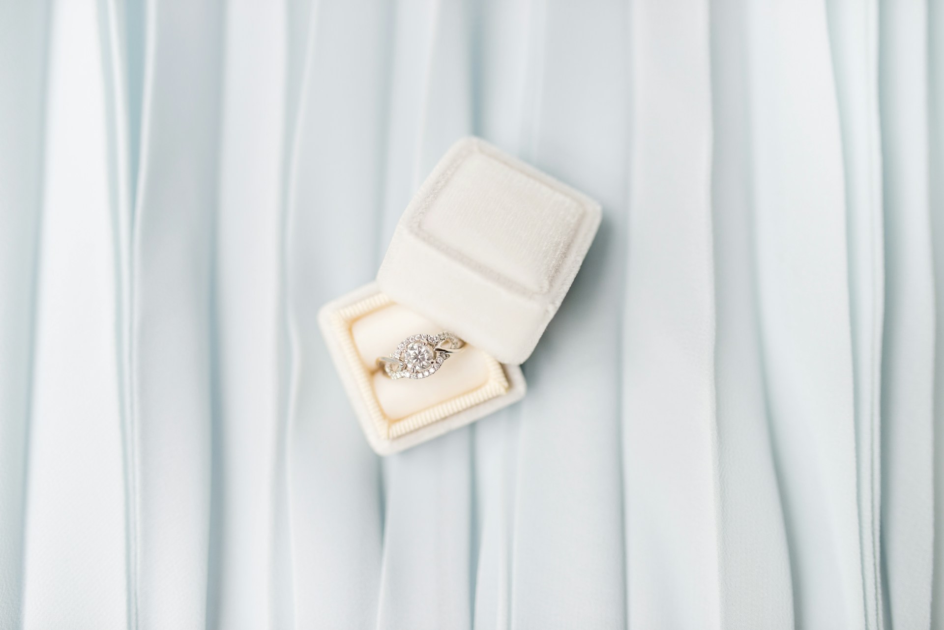 A diamond halo engagement ring in a white box on a white pleated fabric. A diamond halo engagement ring in a white box on a white pleated fabric.