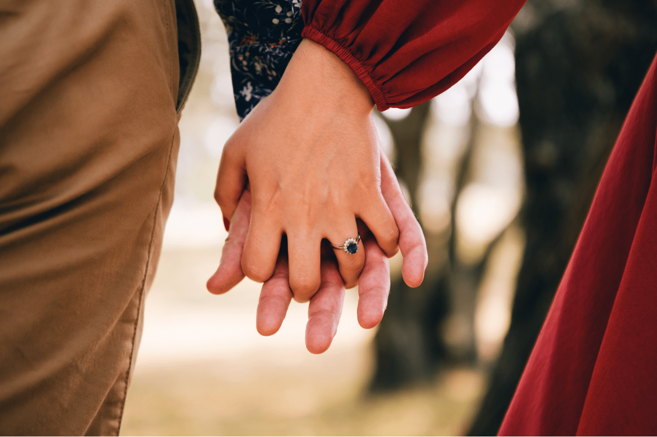 A close-up of a couple holding hands, the woman wearing a sapphire halo engagement ring. A close-up of a couple holding hands, the woman wearing a sapphire halo engagement ring.