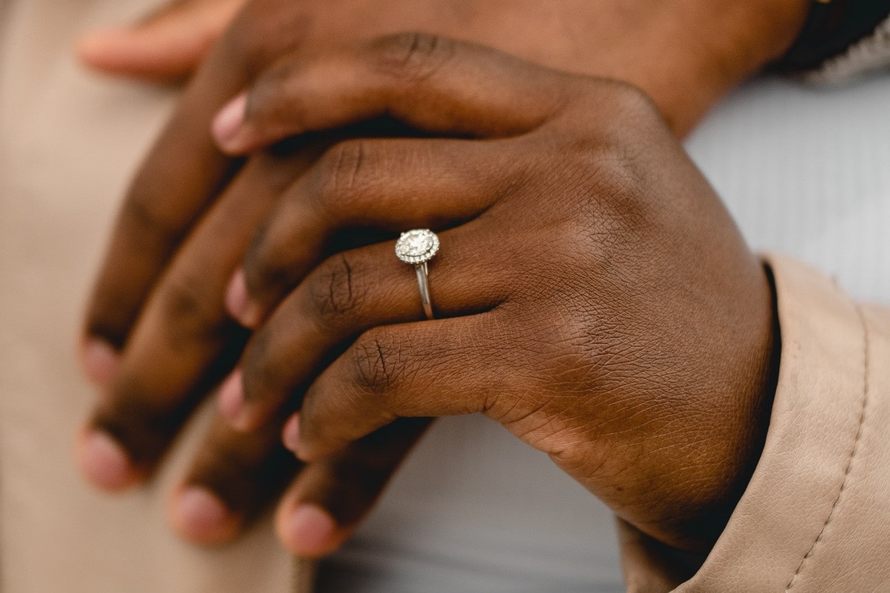 A couple’s hands holding each other, the woman’s hand wearing a silver halo engagement ring. A couple’s hands holding each other, the woman’s hand wearing a silver halo engagement ring.